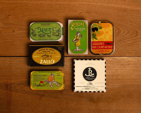 A selection of colourfully-packaged tinned sardines, mackerel and tuna against a wood background.