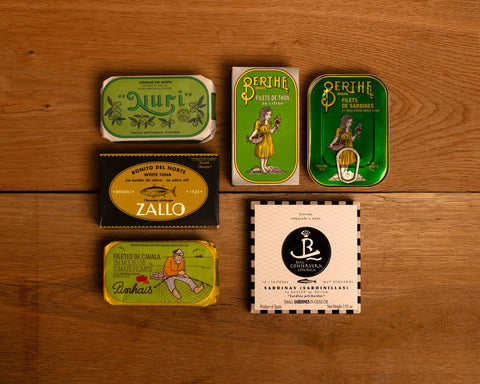 A selection of colourfully-packaged tinned sardines, mackerel and tuna against a wood background.