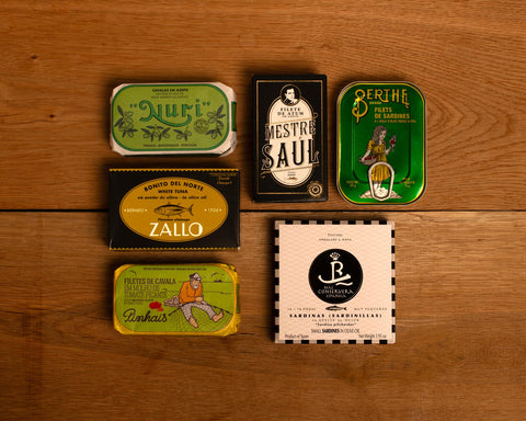 A selection of colourfully-packaged tinned sardines, mackerel and tuna against a wood background.