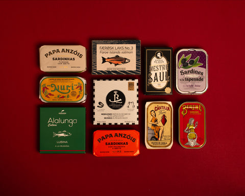 A selection of colourfully-packaged tinned sardines, tuna, mackerel, salmon, mussels and sea bass against a red background.