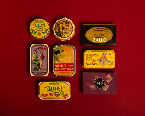 A selection of colourfully-packaged tinned sardines, mackerel, tuna, squid and sardine and mackerel pâtés against a red background.