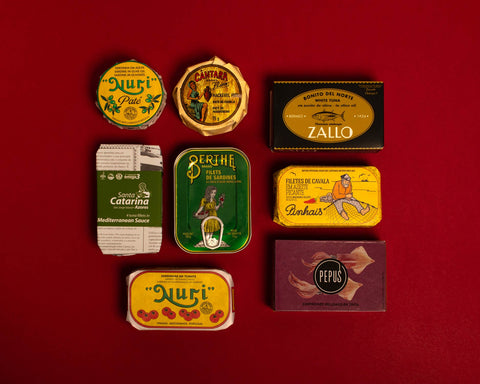 A selection of colourfully-packaged tinned sardines, mackerel, tuna, squid and sardine and mackerel pâtés against a red background.