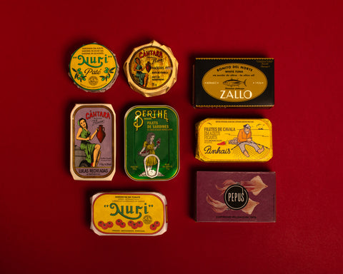 A selection of colourfully-packaged tinned sardines, mackerel, tuna, squid and sardine and mackerel pâtés against a red background.
