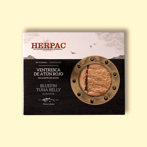 A tin of bluefin tuna belly in rectangular card packaging. The Herpac logo is written in red lettering above an ink-black sea with seagulls flying above it. There is a golden porthole on the right hand side of the packaging through which you can see the glistening bluefin tuna belly.