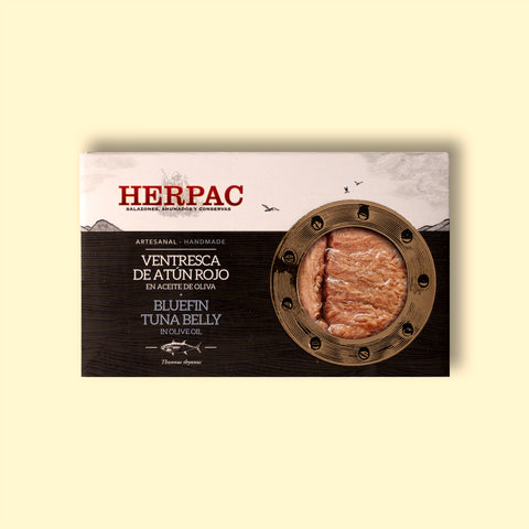 A tin of bluefin tuna belly in rectangular card packaging. The Herpac logo is written in red lettering above an ink-black sea with seagulls flying above it. There is a golden porthole on the right hand side of the packaging through which you can see the glistening bluefin tuna belly.