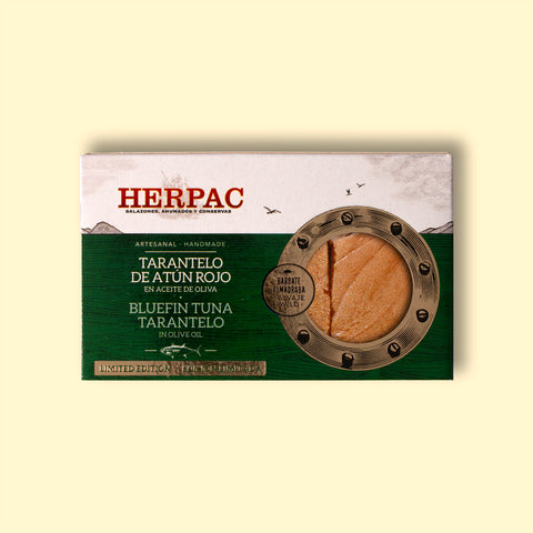 A tin of bluefin tuna tarantelo in rectangular card packaging. The Herpac logo is written in red lettering above an emerald green sea with seagulls flying above it. There is a golden porthole on the right hand side of the packaging through which you can see the glistening tuna tarantelo.