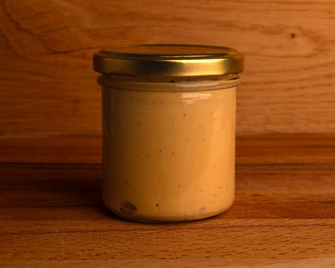 A glass jar of mayonnaise with a golden cap, placed against a wooden background. The jar is transparent, showing the mayonnaise inside. 