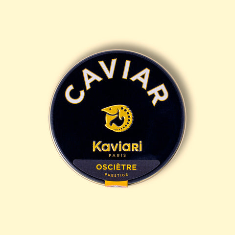 A circular dark blue tin of caviar on a wood backdrop. Caviar is written in white lettering at the top of the tin, above the Kaviari logo: an illustration in gold of a sturgeon fish above Kaviari Paris also in gold lettering. The tin is sealed with a yellow and blue label reading Osciètre prestige.