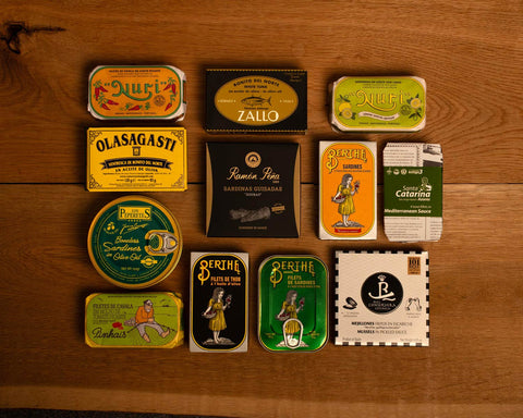 A selection of colourfully-packaged tinned mussels, tuna belly, tuna, sardines and squid against a wood background.