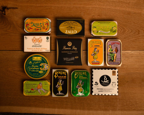 A selection of colourfully-packaged tinned mussels, tuna belly, tuna, sardines and squid against a wood background.