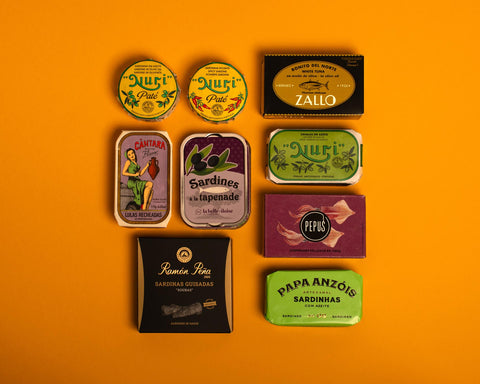 A selection of colourfully-packaged tinned sardines, mackerel, tuna, squid and sardine pâtés against a yellow background.