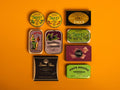 A selection of colourfully-packaged tinned sardines, mackerel, tuna, squid and sardine pâtés against a yellow background.