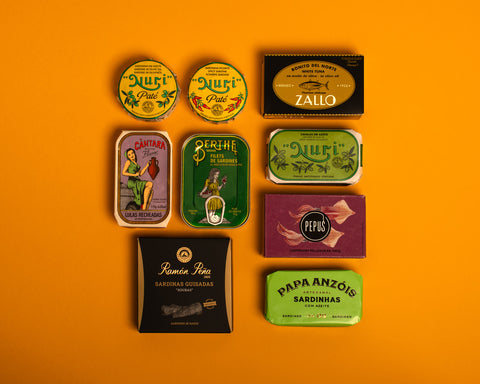 A selection of colourfully-packaged tinned sardines, mackerel, tuna, squid and sardine pâtés against a yellow background.