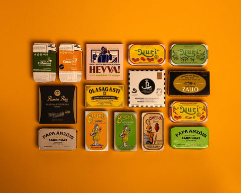 Collection of 15 tins on an orange background. The tins are colourful, some of them have vintage illustrations, some of them are British, some are Spanish.