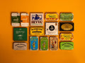Collection of 15 tins on an orange background. The tins are colourful, some of them have vintage illustrations, some of them are British, some are Spanish.