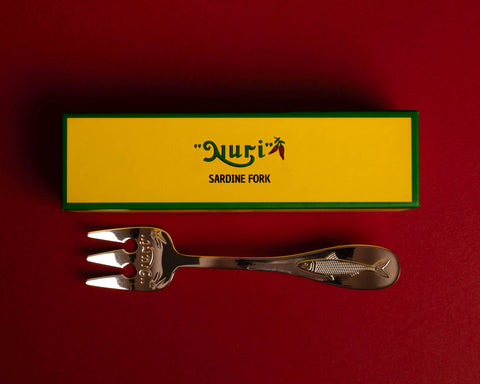 A three-pronged silver sardine fork below a yellow rectangular box with a green border and Nuri logo on it, against a red background.