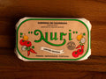 A paper-wrapped tin of Nuri sardines with the Nuri logo in green lettering against beige background, and images of chillies, tomatoes, onion and coriander either side of the log.
