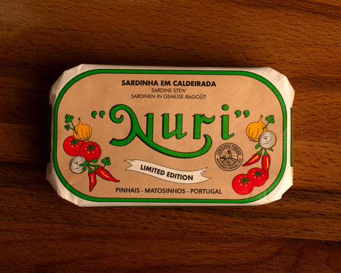 A paper-wrapped tin of Nuri sardines with the Nuri logo in green lettering against beige background, and images of chillies, tomatoes, onion and coriander either side of the log.