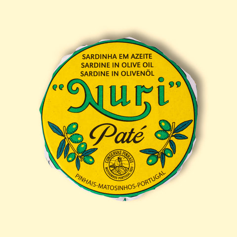 A round tin of sardine pate in a yellow paper wrap. Nuri is written in green lettering above some drawings of olives.