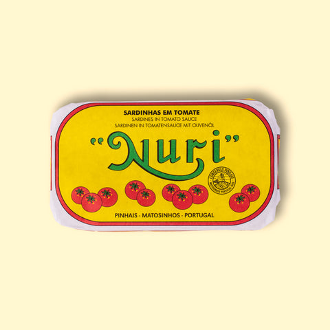 A tin of sardines in a yellow paper wrap with the Nuri logo in green on the front above some images of tomatoes. The tin has a red and white border and is on a wood backdrop.