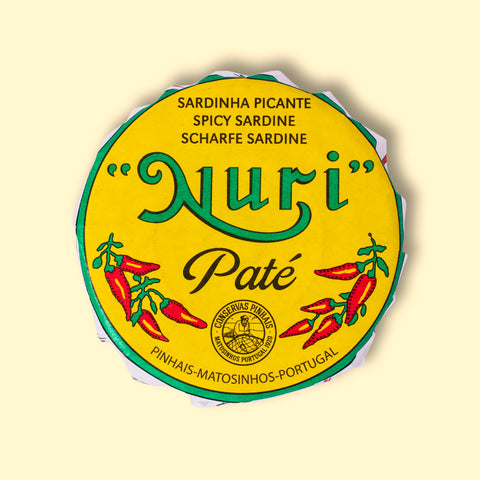 A round tin in a yellow paper wrap. Nuri is written in green lettering above some drawings of red chilli peppers.