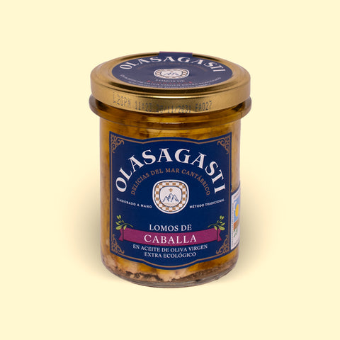 A jar of mackerel fillets in olive oil with a blue label with the Olasagasti arched logo in white lettering on the top, against a wood background.
