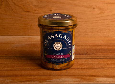 A jar of mackerel fillets in olive oil with a blue label with the Olasagasti arched logo in white lettering on the top, against a wood background.