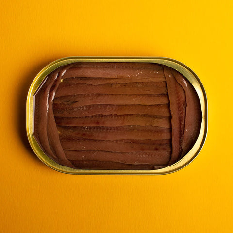 An open gold-coloured tin of anchovy fillets. The fillets are a deep brown and are framed at either side by smaller fillets. Against a rich yellow background.
