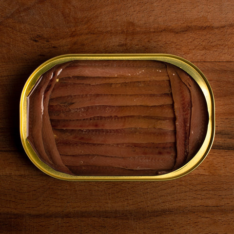 An open gold tin of anchovy fillets. The fillets are a deep brown and are framed at either end by shorter fillets, against a strong dark wooden table