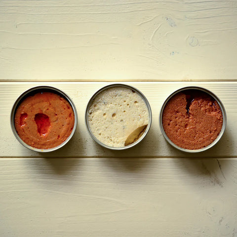 Three open tins of airy-looking pate - one white between two orange-coloured ones - on a pale wood background.