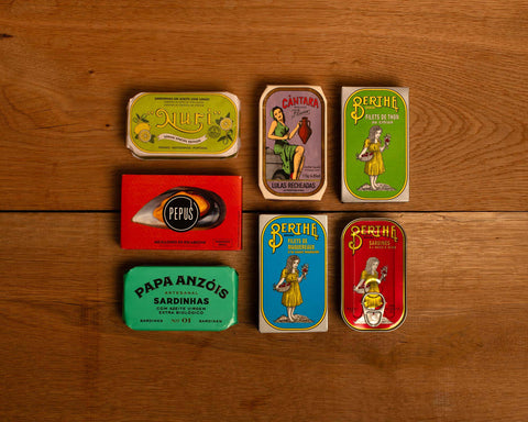 A selection of colourfully-packaged tinned sardines, tuna, squid and mussels against a wood background.