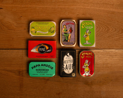 A selection of colourfully-packaged tinned sardines, tuna, squid and mussels against a wood background.
