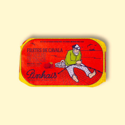 A tin of sardines in a red paper wrap with a yellow border. A fisherman in a green jersey mends his net on the front. The tin is on a wood background.