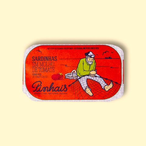 A tin of sardines in a red paper wrap with a white border. A fisherman in a green jersey mends his net on the front. The tin is on a wood background.