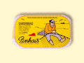 A tin of spicy sardines in a yellow paper wrap with an image of a fisherman mending a fishing net on the front next to a red chilli pepper. The tin is on a wood background.