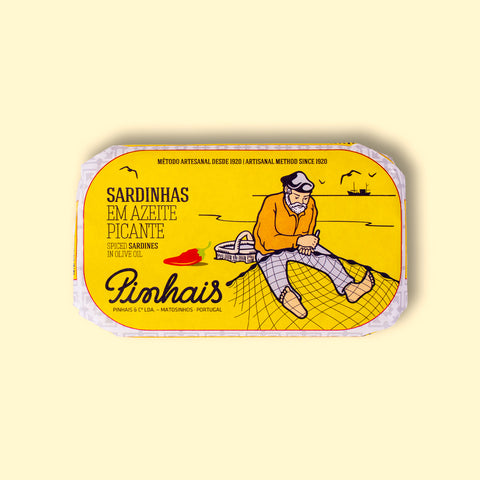 A tin of spicy sardines in a yellow paper wrap with an image of a fisherman mending a fishing net on the front next to a red chilli pepper. The tin is on a wood background.