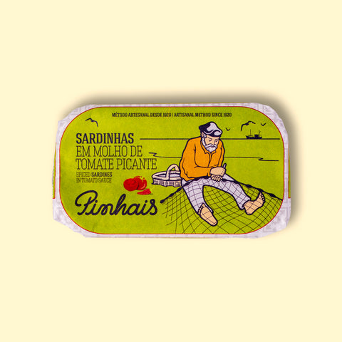 A tin of sardines in a green paper wrap with an image of a fisherman mending a net by some tomatoes and a red chilli pepper. The tin is on a yellow wood backdrop.