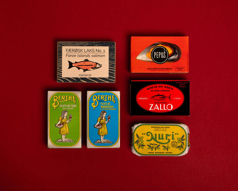 A selection of colourfully-packaged tinned sardines, tuna, mackerel, salmon and carmelo sardines against a red background.