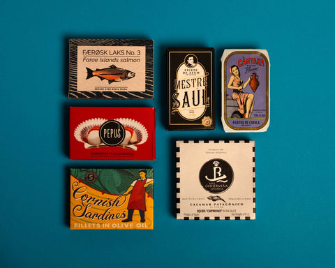 A selection of colourfully-packaged tins of
Cornish sardines, salmon, scallops, mackerel and tuna against a blue background.