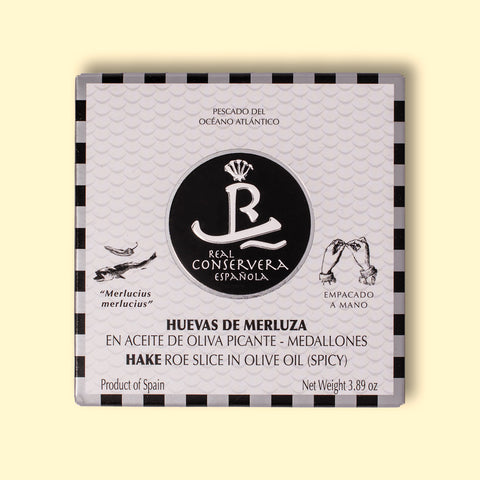 A tin of hake roe slices in olive oil in white packaging, with a silver and black chequered border, featuring an illustration of a hake fish and chilli, against a wooden backdrop.