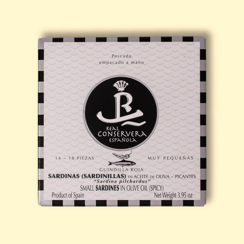 A tin of small spicy sardines in black and white card packaging with a black and silver chequered border, against a wood background.