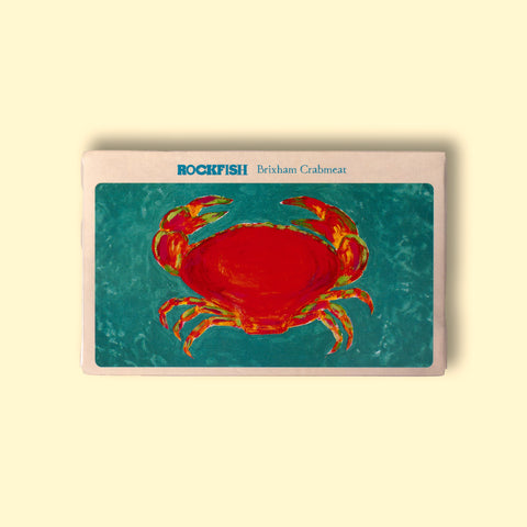 A tin of crab in off-white card packagin with a painting of a vivid orange crab against a clear light blue sea background. The painting is beneath Rockfish Brixham crabmeat written in blue lettering.