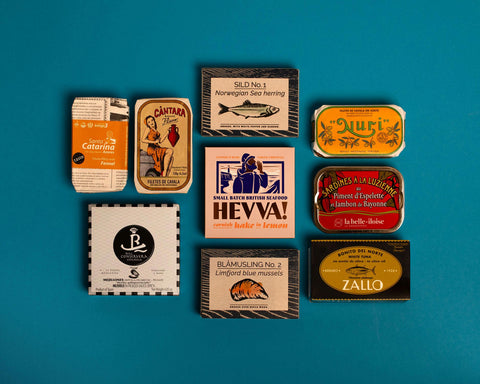 A selection of colourfully-packaged tinned mussels, squid, sardines, herring, tuna and mackerel against a blue background.