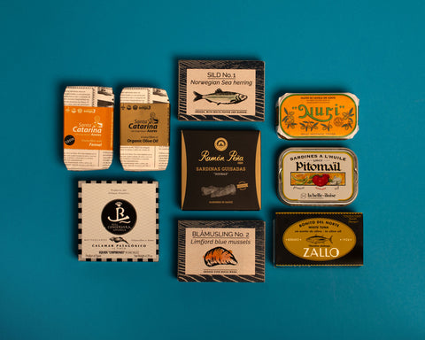 A selection of colourfully-packaged tinned mussels, squid, sardines, herring, tuna and mackerel against a blue background.
