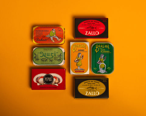 A selection of colourfully-packaged tinned scallops, tuna, mackerel and sardines against a yellow background.