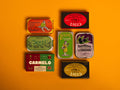 A selection of colourfully-packaged tinned scallops, tuna, mackerel and sardines against a yellow background.