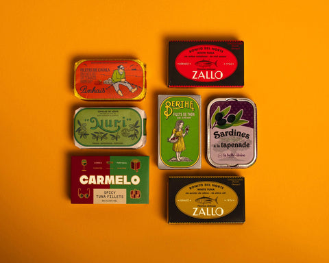 A selection of colourfully-packaged tinned scallops, tuna, mackerel and sardines against a yellow background.