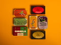 A selection of colourfully-packaged tinned scallops, tuna, mackerel and sardines against a yellow background.