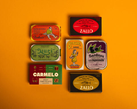 A selection of colourfully-packaged tinned scallops, tuna, mackerel and sardines against a yellow background.
