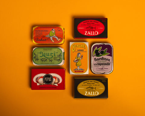 A selection of colourfully-packaged tinned scallops, tuna, mackerel and sardines against a yellow background.
